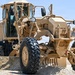 A U.S. Air Force motor grader painted in desert tan/sand color is photographed at close range on a gravel or sandy terrain. The vehicle bears a U.S. Air Force registration plate reading '21D 00054'. It appears to be engaged in road grading or construction operations in an arid environment.