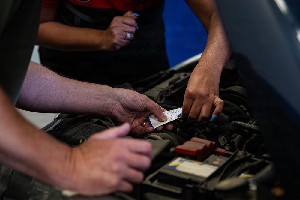 Two or more people are working together over an open car engine bay, with one person holding a small white card or label near the engine components. The scene appears to take place in an automotive garage or workshop setting. Multiple hands are visible, suggesting a collaborative inspection or repair task.