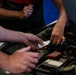 Two or more people are working together over an open car engine bay, with one person holding a small white card or label near the engine components. The scene appears to take place in an automotive garage or workshop setting. Multiple hands are visible, suggesting a collaborative inspection or repair task.