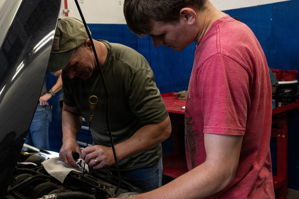 Two individuals are working together on a vehicle engine in what appears to be an automotive garage or repair shop. An older man wearing a green cap and olive t-shirt is leaning over the engine bay while a younger man in a red t-shirt observes or assists. A third person is partially visible in the background.