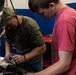 Two individuals are working together on a vehicle engine in what appears to be an automotive garage or repair shop. An older man wearing a green cap and olive t-shirt is leaning over the engine bay while a younger man in a red t-shirt observes or assists. A third person is partially visible in the background.