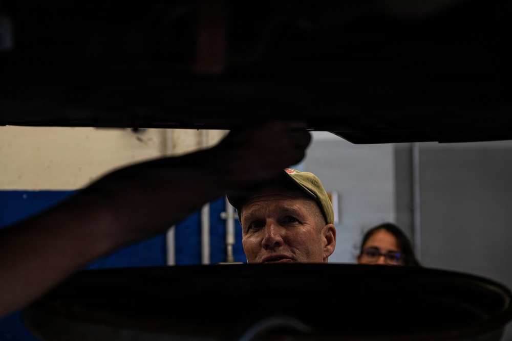 A man wearing a baseball cap is looking upward at the underside of a raised vehicle, likely in an automotive repair shop or garage. A second person, appearing to be a woman wearing glasses, is visible in the background. The perspective is shot from below the vehicle, creating a dramatic low-angle composition.