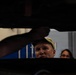 A man wearing a baseball cap is looking upward at the underside of a raised vehicle, likely in an automotive repair shop or garage. A second person, appearing to be a woman wearing glasses, is visible in the background. The perspective is shot from below the vehicle, creating a dramatic low-angle composition.