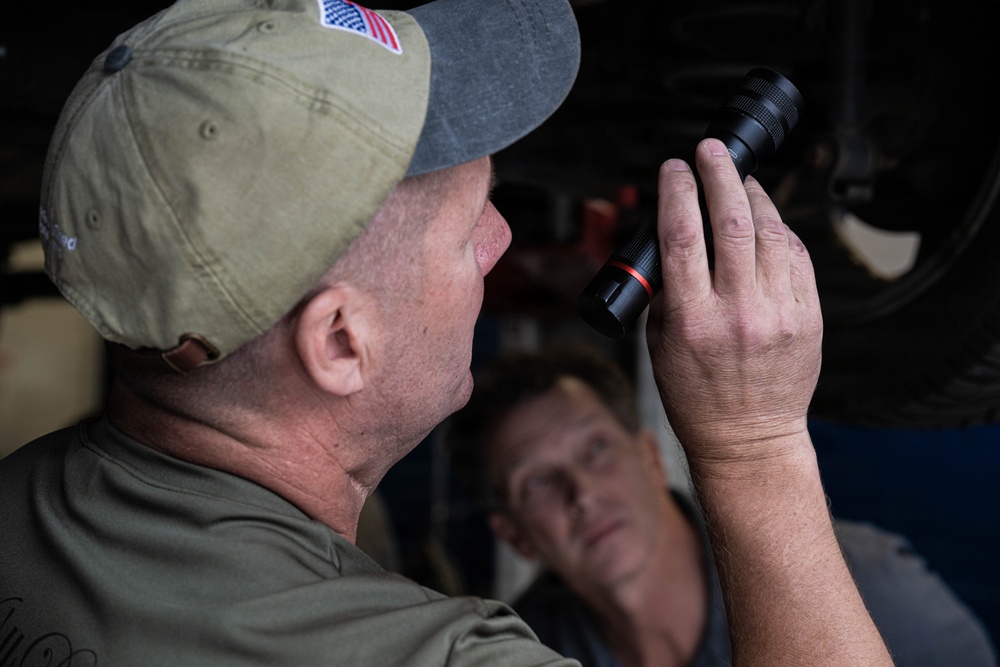 Two men are visible in what appears to be an automotive garage or repair shop setting. The foreground figure, wearing an olive green cap with an American flag patch, is holding and examining a black tactical flashlight with a red accent ring. A second man is visible in the background, slightly out of focus, observing the scene.