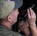 Two men are visible in what appears to be an automotive garage or repair shop setting. The foreground figure, wearing an olive green cap with an American flag patch, is holding and examining a black tactical flashlight with a red accent ring. A second man is visible in the background, slightly out of focus, observing the scene.