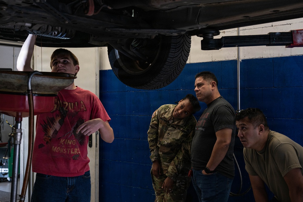 A mechanic in a red Godzilla t-shirt is explaining or demonstrating something about the undercarriage of a vehicle raised on a lift to three observers. Two of the observers appear to be in military uniform or military-style clothing, and all three are leaning in to get a closer look at the underside of the vehicle. The scene takes place in an automotive repair shop or garage.