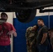 A mechanic in a red Godzilla t-shirt is explaining or demonstrating something about the undercarriage of a vehicle raised on a lift to three observers. Two of the observers appear to be in military uniform or military-style clothing, and all three are leaning in to get a closer look at the underside of the vehicle. The scene takes place in an automotive repair shop or garage.