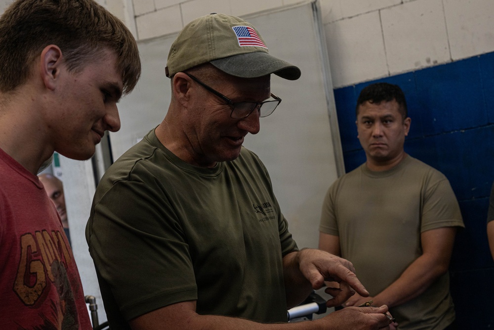 An older man wearing a US flag baseball cap, glasses, and an olive green t-shirt appears to be instructing or demonstrating something to a younger man in a red t-shirt. A third individual in a tan/khaki t-shirt observes in the background. The setting appears to be an indoor facility with painted cinder block walls.