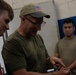 An older man wearing a US flag baseball cap, glasses, and an olive green t-shirt appears to be instructing or demonstrating something to a younger man in a red t-shirt. A third individual in a tan/khaki t-shirt observes in the background. The setting appears to be an indoor facility with painted cinder block walls.