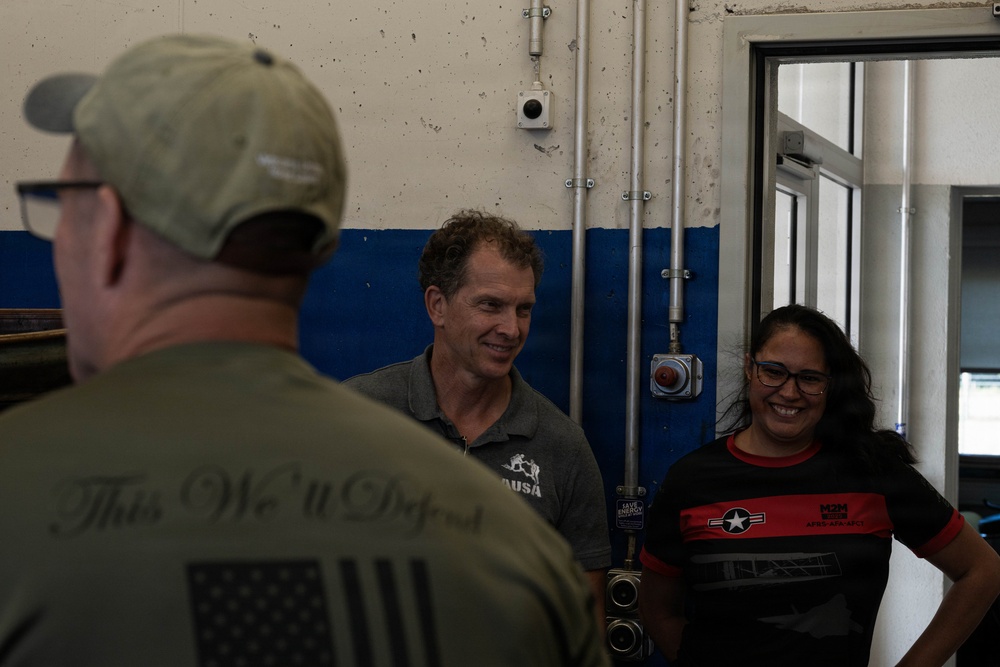 Three individuals are engaged in conversation inside what appears to be an industrial or military facility. The person in the foreground faces away from the camera, wearing a shirt with 'This We'll Defend' and an American flag motif. The other two individuals — a middle-aged man in a gray polo shirt with an 'AUSA' logo and a woman in a black and red shirt — are smiling and appear engaged in a friendly exchange.