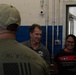 Three individuals are engaged in conversation inside what appears to be an industrial or military facility. The person in the foreground faces away from the camera, wearing a shirt with 'This We'll Defend' and an American flag motif. The other two individuals — a middle-aged man in a gray polo shirt with an 'AUSA' logo and a woman in a black and red shirt — are smiling and appear engaged in a friendly exchange.