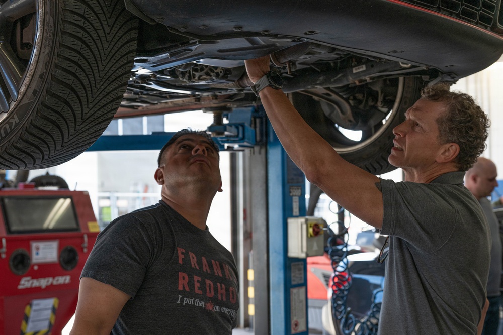 Two men are examining the underside of a vehicle that has been raised on a hydraulic lift in an automotive repair shop. One man is pointing at or touching a component beneath the car while the other looks upward to observe. The setting is a professional garage environment with visible tools and equipment.