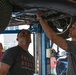 Two men are examining the underside of a vehicle that has been raised on a hydraulic lift in an automotive repair shop. One man is pointing at or touching a component beneath the car while the other looks upward to observe. The setting is a professional garage environment with visible tools and equipment.