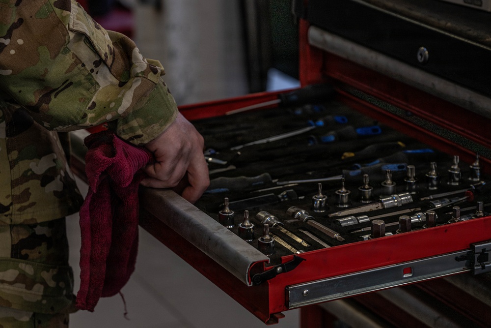 A person wearing a military multicam/OCP camouflage uniform is accessing an open red tool chest drawer containing various mechanical tools. The individual is holding a red shop rag in their hand while gripping the drawer edge. The image appears to depict a military maintenance or repair environment.