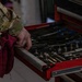 A person wearing a military multicam/OCP camouflage uniform is accessing an open red tool chest drawer containing various mechanical tools. The individual is holding a red shop rag in their hand while gripping the drawer edge. The image appears to depict a military maintenance or repair environment.