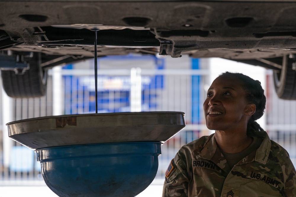A U.S. Army soldier named Brown, wearing OCP (Operational Camouflage Pattern) uniform, is performing vehicle maintenance underneath a raised vehicle. She is smiling while observing oil draining from the vehicle's undercarriage into a collection pan. The setting appears to be a military or commercial automotive service facility.