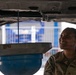 A U.S. Army soldier named Brown, wearing OCP (Operational Camouflage Pattern) uniform, is performing vehicle maintenance underneath a raised vehicle. She is smiling while observing oil draining from the vehicle's undercarriage into a collection pan. The setting appears to be a military or commercial automotive service facility.