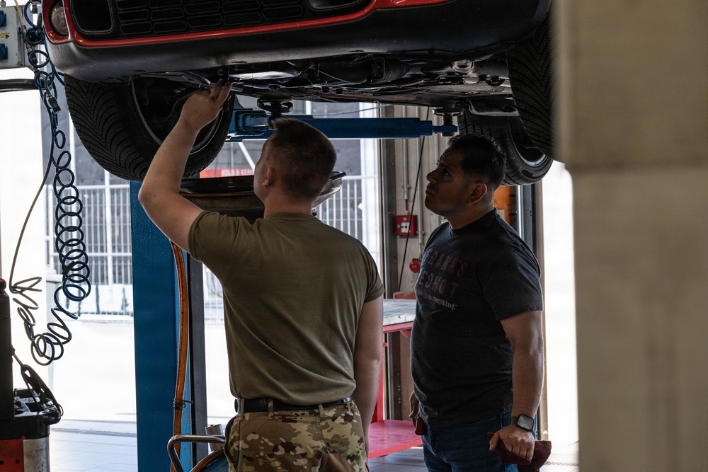 Two men are working in an automotive repair bay where a red vehicle is raised on a hydraulic lift. One man wearing military camouflage (MultiCam) pants and an olive t-shirt is reaching up to work on the undercarriage of the vehicle, while a second man in a dark graphic t-shirt and jeans observes. The setting appears to be a professional or military auto shop facility.