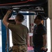 Two men are working in an automotive repair bay where a red vehicle is raised on a hydraulic lift. One man wearing military camouflage (MultiCam) pants and an olive t-shirt is reaching up to work on the undercarriage of the vehicle, while a second man in a dark graphic t-shirt and jeans observes. The setting appears to be a professional or military auto shop facility.