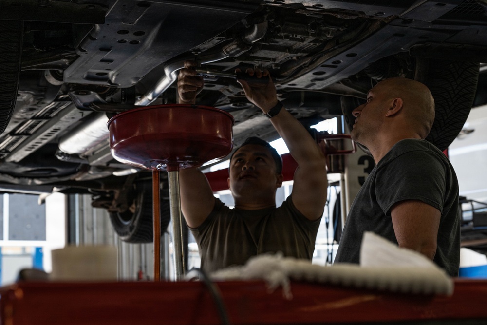 Two mechanics are working underneath a raised vehicle in an automotive repair shop. One mechanic is actively using tools on the undercarriage while the other observes closely, suggesting a training or collaborative maintenance scenario. A red oil drain pan on a stand is positioned to catch fluids during the service procedure.