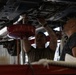 Two mechanics are working underneath a raised vehicle in an automotive repair shop. One mechanic is actively using tools on the undercarriage while the other observes closely, suggesting a training or collaborative maintenance scenario. A red oil drain pan on a stand is positioned to catch fluids during the service procedure.