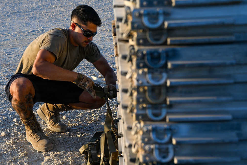 A young male in military-style attire is crouched down working on a large stacked metal equipment rack or pallet system, likely in an arid outdoor environment. He appears to be securing or adjusting straps and fasteners on the equipment. The setting and gear suggest a military logistics or equipment maintenance context.