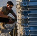 A young male in military-style attire is crouched down working on a large stacked metal equipment rack or pallet system, likely in an arid outdoor environment. He appears to be securing or adjusting straps and fasteners on the equipment. The setting and gear suggest a military logistics or equipment maintenance context.