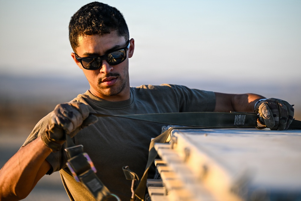 A young male in military-style attire is working with straps and equipment in an outdoor setting with warm, golden-hour lighting. He is wearing tactical gloves, an olive drab t-shirt, and dark wraparound sunglasses. He appears to be securing or adjusting cargo straps on a piece of equipment or vehicle.