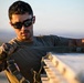 A young male in military-style attire is working with straps and equipment in an outdoor setting with warm, golden-hour lighting. He is wearing tactical gloves, an olive drab t-shirt, and dark wraparound sunglasses. He appears to be securing or adjusting cargo straps on a piece of equipment or vehicle.