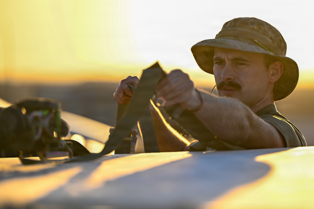 A man wearing a tan/olive boonie hat and mustache is handling dark straps or harness equipment, likely securing or adjusting gear on a vehicle or surface. The scene is bathed in warm golden sunset light, creating a dramatic backlit atmosphere. The setting appears to be an open, arid or flat outdoor environment.