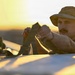 A man wearing a tan/olive boonie hat and mustache is handling dark straps or harness equipment, likely securing or adjusting gear on a vehicle or surface. The scene is bathed in warm golden sunset light, creating a dramatic backlit atmosphere. The setting appears to be an open, arid or flat outdoor environment.