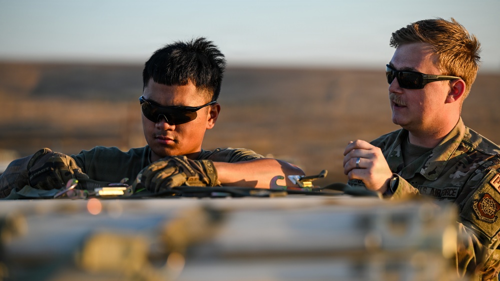Two U.S. Air Force personnel in OCP (Operational Camouflage Pattern) uniforms are working together on equipment in what appears to be an arid, desert-like environment. The image is taken during golden hour lighting, suggesting early morning or late afternoon. One airman appears to be performing a hands-on task while the other observes or provides guidance.
