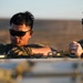 Two U.S. Air Force personnel in OCP (Operational Camouflage Pattern) uniforms are working together on equipment in what appears to be an arid, desert-like environment. The image is taken during golden hour lighting, suggesting early morning or late afternoon. One airman appears to be performing a hands-on task while the other observes or provides guidance.