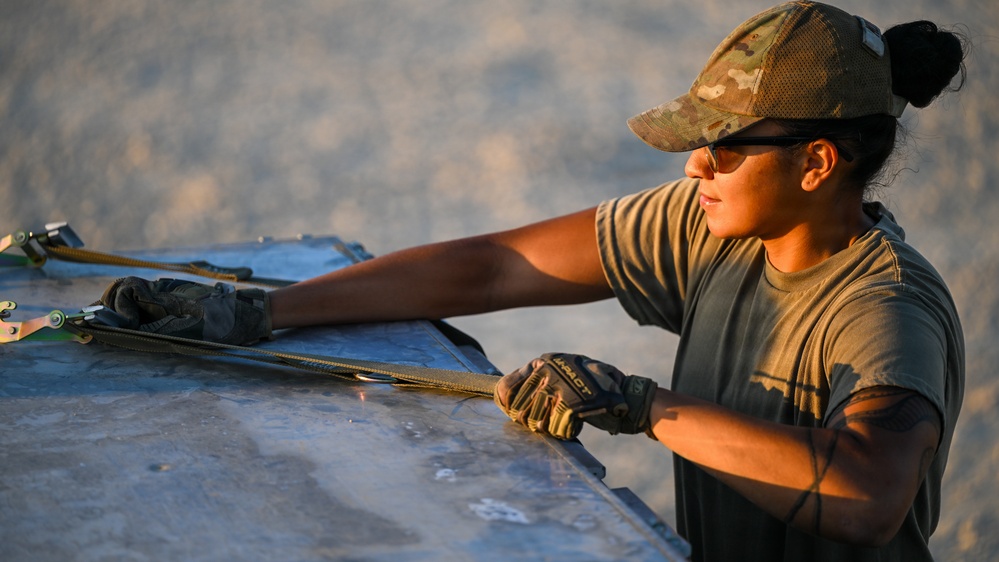 A female military service member wearing OCP (Operational Camouflage Pattern) uniform and cap is working with cargo straps or tie-downs on what appears to be a metal surface, likely a pallet or equipment panel. She is wearing tactical gloves branded 'Mechanix' and sunglasses, working in what appears to be a desert or arid environment during golden hour lighting. The image captures her focused on securing or adjusting load restraint equipment.