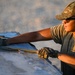 A female military service member wearing OCP (Operational Camouflage Pattern) uniform and cap is working with cargo straps or tie-downs on what appears to be a metal surface, likely a pallet or equipment panel. She is wearing tactical gloves branded 'Mechanix' and sunglasses, working in what appears to be a desert or arid environment during golden hour lighting. The image captures her focused on securing or adjusting load restraint equipment.