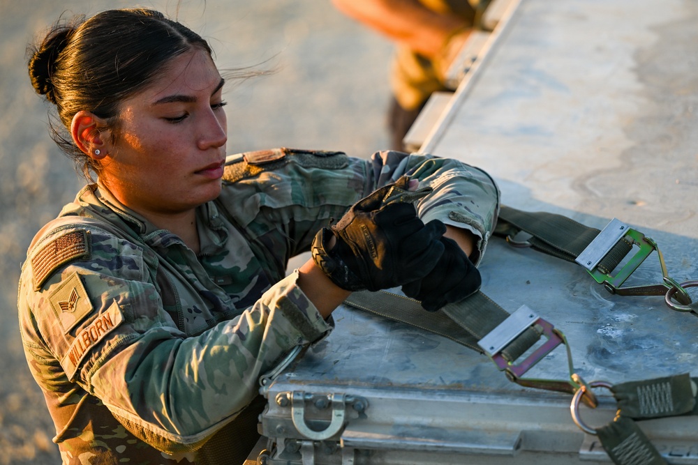 A female U.S. Air Force service member, identified by name tape as 'Willborn,' is working with straps and hardware on a metal equipment case or pallet. She is wearing OCP (Operational Camouflage Pattern) uniform with tactical gloves and has the rank insignia of a Senior Airman visible on her chest. The image is taken during golden hour lighting, suggesting early morning or late afternoon operations.