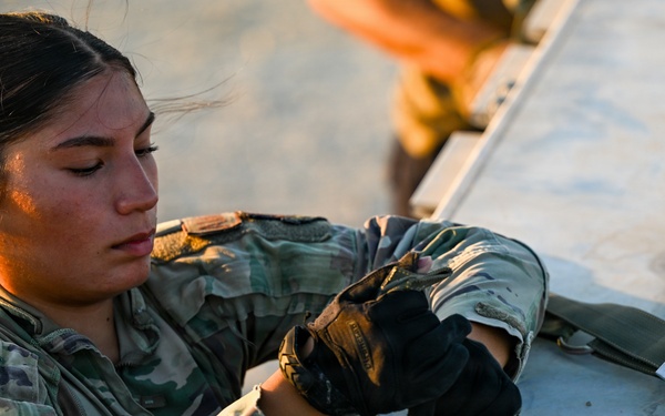 A female U.S. Air Force service member, identified by name tape as 'Willborn,' is working with straps and hardware on a metal equipment case or pallet. She is wearing OCP (Operational Camouflage Pattern) uniform with tactical gloves and has the rank insignia of a Senior Airman visible on her chest. The image is taken during golden hour lighting, suggesting early morning or late afternoon operations.