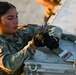 A female U.S. Air Force service member, identified by name tape as 'Willborn,' is working with straps and hardware on a metal equipment case or pallet. She is wearing OCP (Operational Camouflage Pattern) uniform with tactical gloves and has the rank insignia of a Senior Airman visible on her chest. The image is taken during golden hour lighting, suggesting early morning or late afternoon operations.