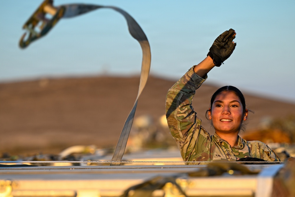 A female military service member in MultiCam/OCP camouflage uniform is working atop a vehicle or platform, tossing or releasing a ratchet strap into the air. She is wearing black tactical gloves and smiling, suggesting a routine work task being performed in good spirits. The setting appears to be an arid, desert-like environment during golden hour lighting.