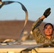 A female military service member in MultiCam/OCP camouflage uniform is working atop a vehicle or platform, tossing or releasing a ratchet strap into the air. She is wearing black tactical gloves and smiling, suggesting a routine work task being performed in good spirits. The setting appears to be an arid, desert-like environment during golden hour lighting.