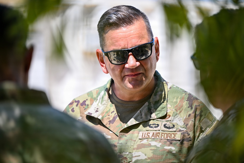 A U.S. Air Force enlisted member wearing Operational Camouflage Pattern (OCP) uniform is photographed in an outdoor setting with foliage in the foreground. The individual wears dark sunglasses with blue frames and displays multiple badges on their uniform. The image appears to be taken in a bright, sunny environment, possibly an overseas or warm-climate location.