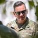 A U.S. Air Force enlisted member wearing Operational Camouflage Pattern (OCP) uniform is photographed in an outdoor setting with foliage in the foreground. The individual wears dark sunglasses with blue frames and displays multiple badges on their uniform. The image appears to be taken in a bright, sunny environment, possibly an overseas or warm-climate location.