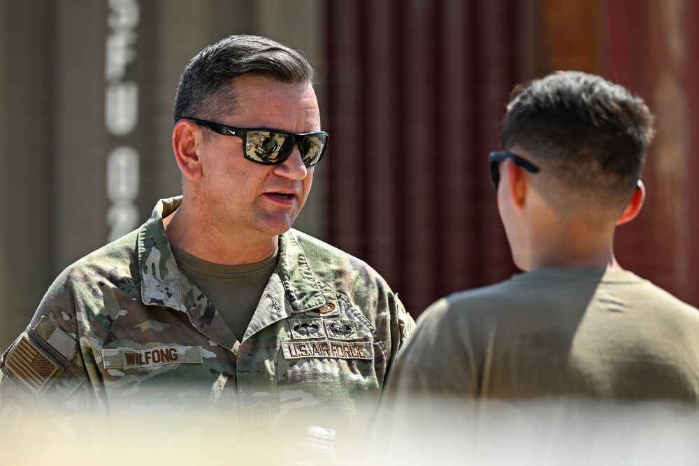 Two U.S. Air Force personnel are engaged in a conversation outdoors near what appear to be shipping containers. The individual facing the camera wears OCP (Operational Camouflage Pattern) uniform with a name tape reading 'WILFONG' and a 'U.S. AIR FORCE' branch tape, along with visible qualification badges. Both individuals are wearing sunglasses, suggesting bright outdoor conditions.