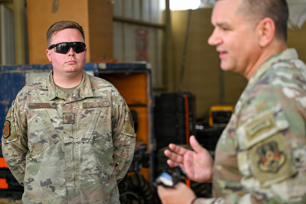Two U.S. Air Force personnel in OCP (Operational Camouflage Pattern) uniforms are engaged in conversation inside what appears to be a maintenance hangar or storage facility. The airman on the left, whose name tape reads 'TRONNIER' and wears Staff Sergeant rank insignia, is listening attentively while wearing tactical sunglasses. The individual on the right, partially out of focus, appears to be speaking and gesturing with his hands.