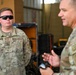 Two U.S. Air Force personnel in OCP (Operational Camouflage Pattern) uniforms are engaged in conversation inside what appears to be a maintenance hangar or storage facility. The airman on the left, whose name tape reads 'TRONNIER' and wears Staff Sergeant rank insignia, is listening attentively while wearing tactical sunglasses. The individual on the right, partially out of focus, appears to be speaking and gesturing with his hands.