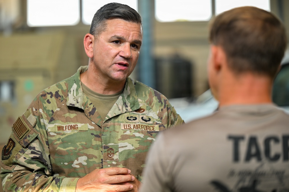 A U.S. Air Force senior enlisted member named Wilfong, wearing OCP (Operational Camouflage Pattern) uniform, is engaged in conversation with another individual wearing a tan tactical t-shirt. The uniformed individual displays a Chief Master Sergeant rank insignia along with airborne and other qualification badges. The setting appears to be an indoor military facility or hangar.