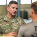 A U.S. Air Force senior enlisted member named Wilfong, wearing OCP (Operational Camouflage Pattern) uniform, is engaged in conversation with another individual wearing a tan tactical t-shirt. The uniformed individual displays a Chief Master Sergeant rank insignia along with airborne and other qualification badges. The setting appears to be an indoor military facility or hangar.