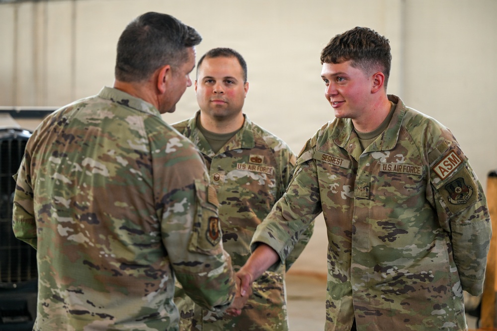 Three U.S. Air Force personnel in OCP (Operational Camouflage Pattern) uniforms are engaged in a handshake interaction, likely a formal recognition or greeting ceremony. The airman on the right, identified by the name tape 'SEGREST,' is shaking hands with a senior individual whose back is to the camera, while a third airman observes from the background. The setting appears to be inside a military hangar or facility.