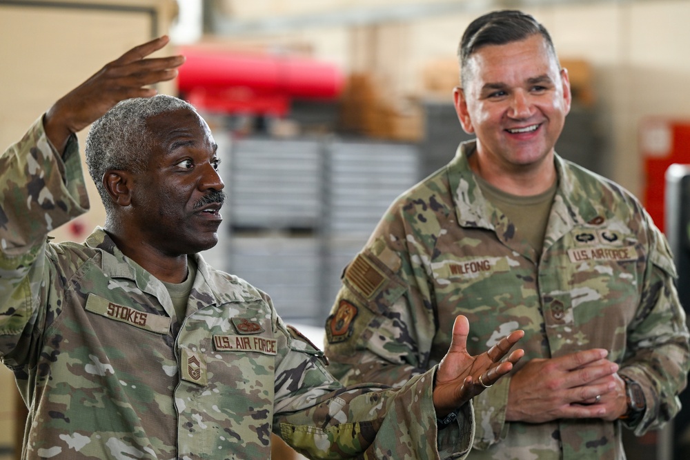 Two U.S. Air Force personnel in OCP (Operational Camouflage Pattern) uniforms are engaged in an animated conversation inside what appears to be a military facility or hangar. The individual on the left, identified by name tape as 'STOKES,' is gesturing expressively while speaking, and the individual on the right, identified as 'WILFONG,' is smiling and listening. Both appear to be senior non-commissioned officers based on their rank insignia.