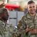 Two U.S. Air Force personnel in OCP (Operational Camouflage Pattern) uniforms are engaged in an animated conversation inside what appears to be a military facility or hangar. The individual on the left, identified by name tape as 'STOKES,' is gesturing expressively while speaking, and the individual on the right, identified as 'WILFONG,' is smiling and listening. Both appear to be senior non-commissioned officers based on their rank insignia.